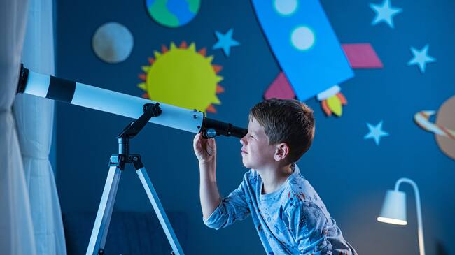 Curious boy using telescope to explore moon surface at night in his bedroom. Young child using telescope to see remote galaxy from room with decorated wall with rocket, planets, stars and cardboard solar system. Passionate kid in pajamas looking at stars using telescope, astronomy and stargazing concept.