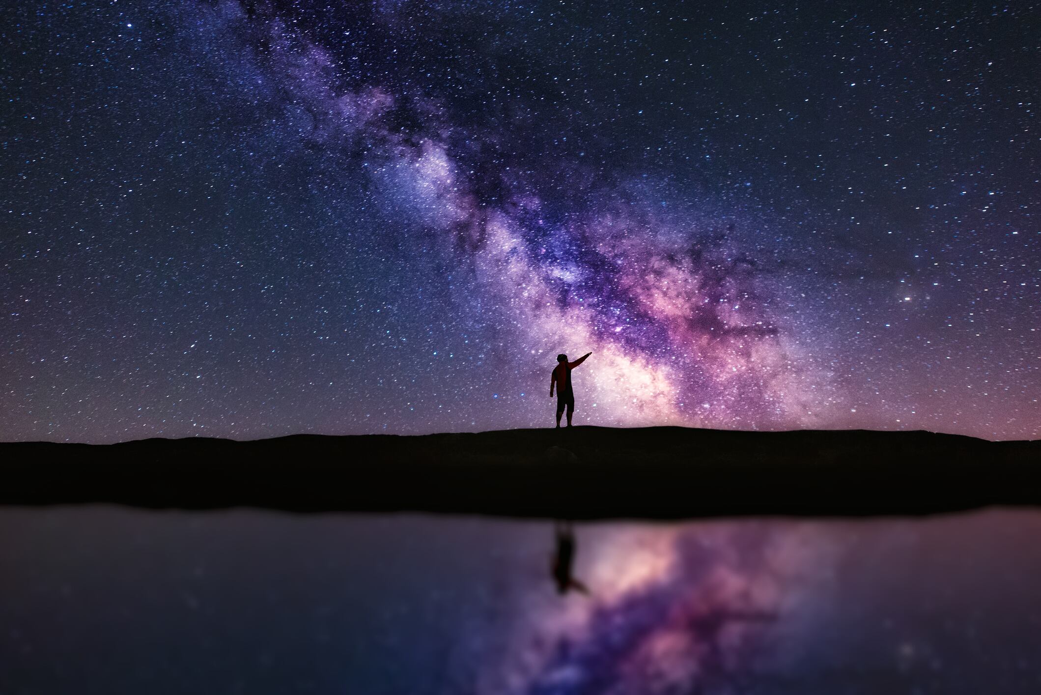 Person pointing up the Milky Way galaxy at night. The scene is reflected in water. A Veiga, Ourense, Spain