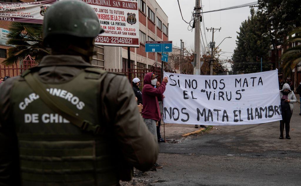 A policeman looks at people holding a sign reading If the virus does not kill us, hunger does to protest for not getting aid from the government amid the lockdown imposed against the spread of the new coronavirus at a low-income area in La Pintana commune, Santiago, on May 20, 2020. (Photo by Martin BERNETTI / AFP) (Photo by MARTIN BERNETTI/AFP via Getty Images)