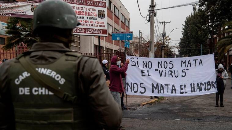 A policeman looks at people holding a sign reading If the virus does not kill us, hunger does to protest for not getting aid from the government amid the lockdown imposed against the spread of the new coronavirus at a low-income area in La Pintana commune, Santiago, on May 20, 2020. (Photo by Martin BERNETTI / AFP) (Photo by MARTIN BERNETTI/AFP via Getty Images)