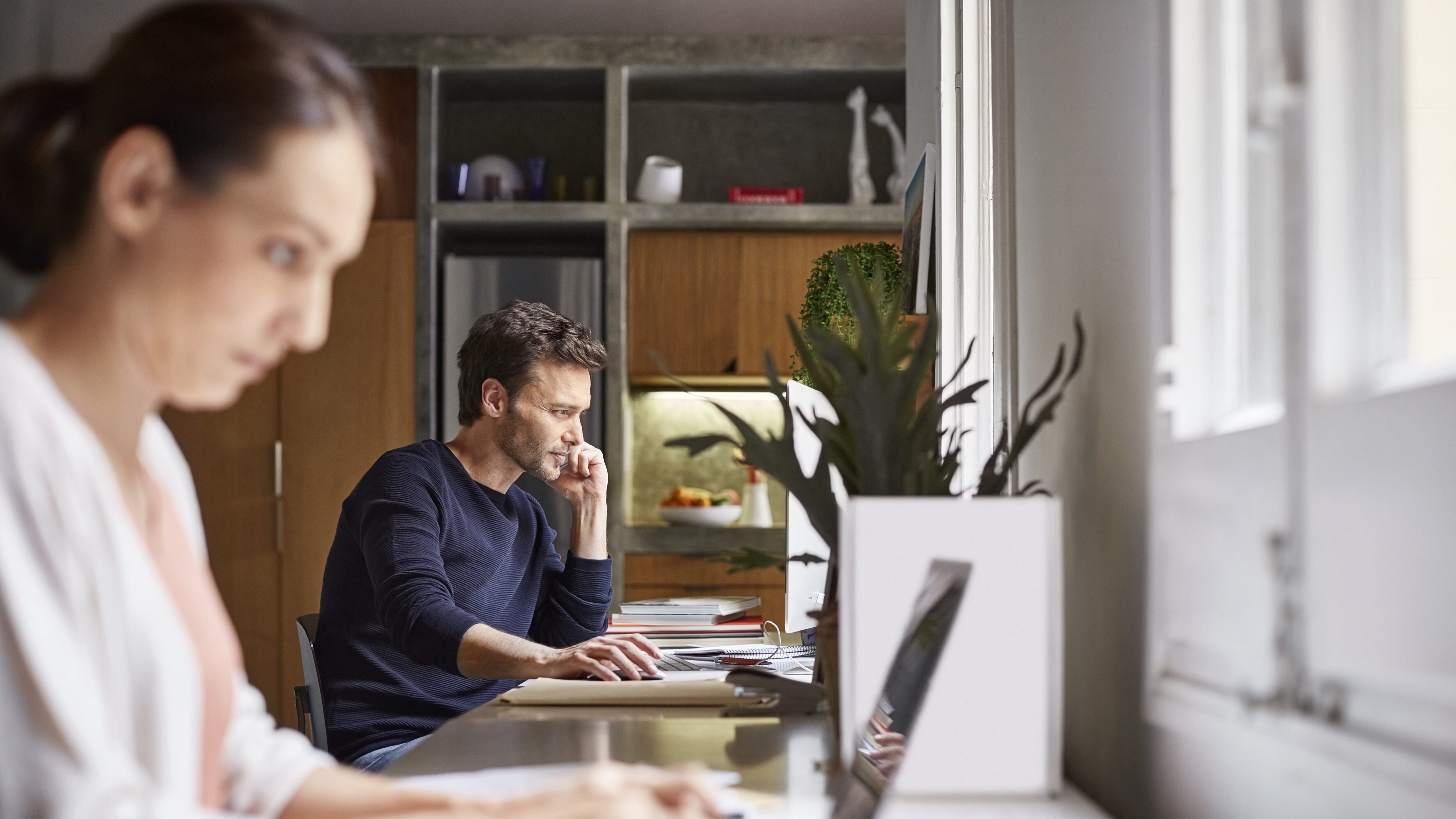 Mid adult man using computer. Woman is working in foreground at desk. Business couple are in home office.