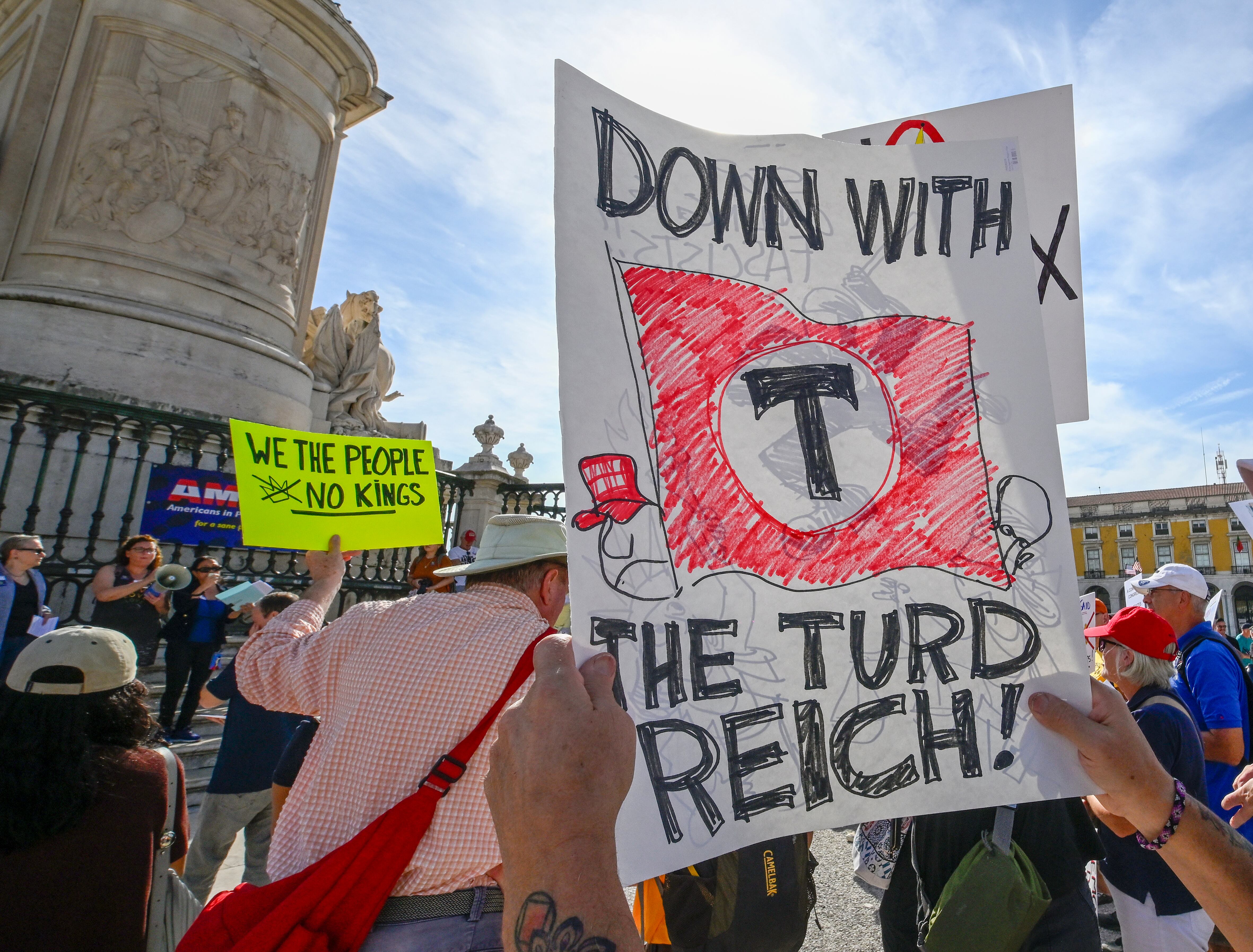 LISBON, PORTUGAL - OCTOBER 18: (Editors note: Image contains profanity) U.S. nationals residing in Portugal hold placards and chant while gathering in Praça do Comercio at a protest organized by American activists living in Portugal, during a "No Kings" protest against U.S. President Donald Trump on October 18, 2025 in Lisbon, Portugal. The protest follows similar "No Kings" events in the United States. (Photo by Horacio Villalobos#Corbis/Getty Images)