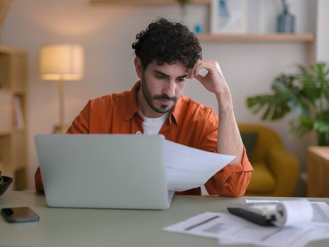 View of a young man, sitting in his living room at home next to his laptop, with worried expression while checking home bills. Concept of home economics.