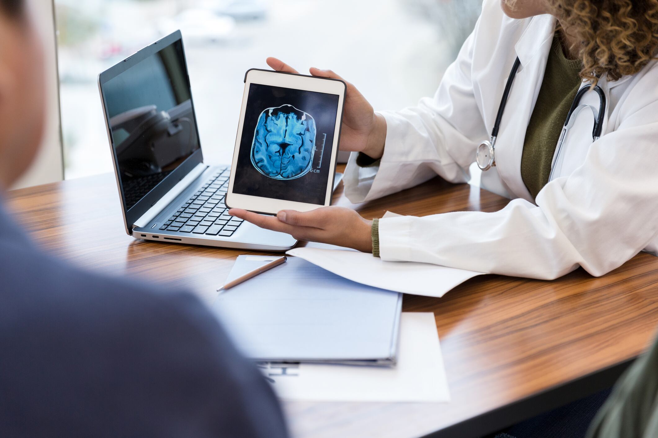Unrecognizable female doctor shows a male patient an image of his brain from an MRI scan. The image is on a digital tablet.