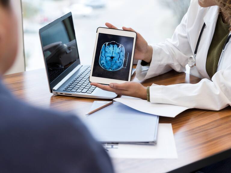 Unrecognizable female doctor shows a male patient an image of his brain from an MRI scan. The image is on a digital tablet.