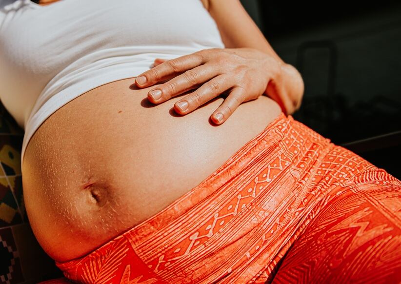 A pregnant woman sits comfortably outdoors, resting her hand on her belly. Focus is on her growing bump.
