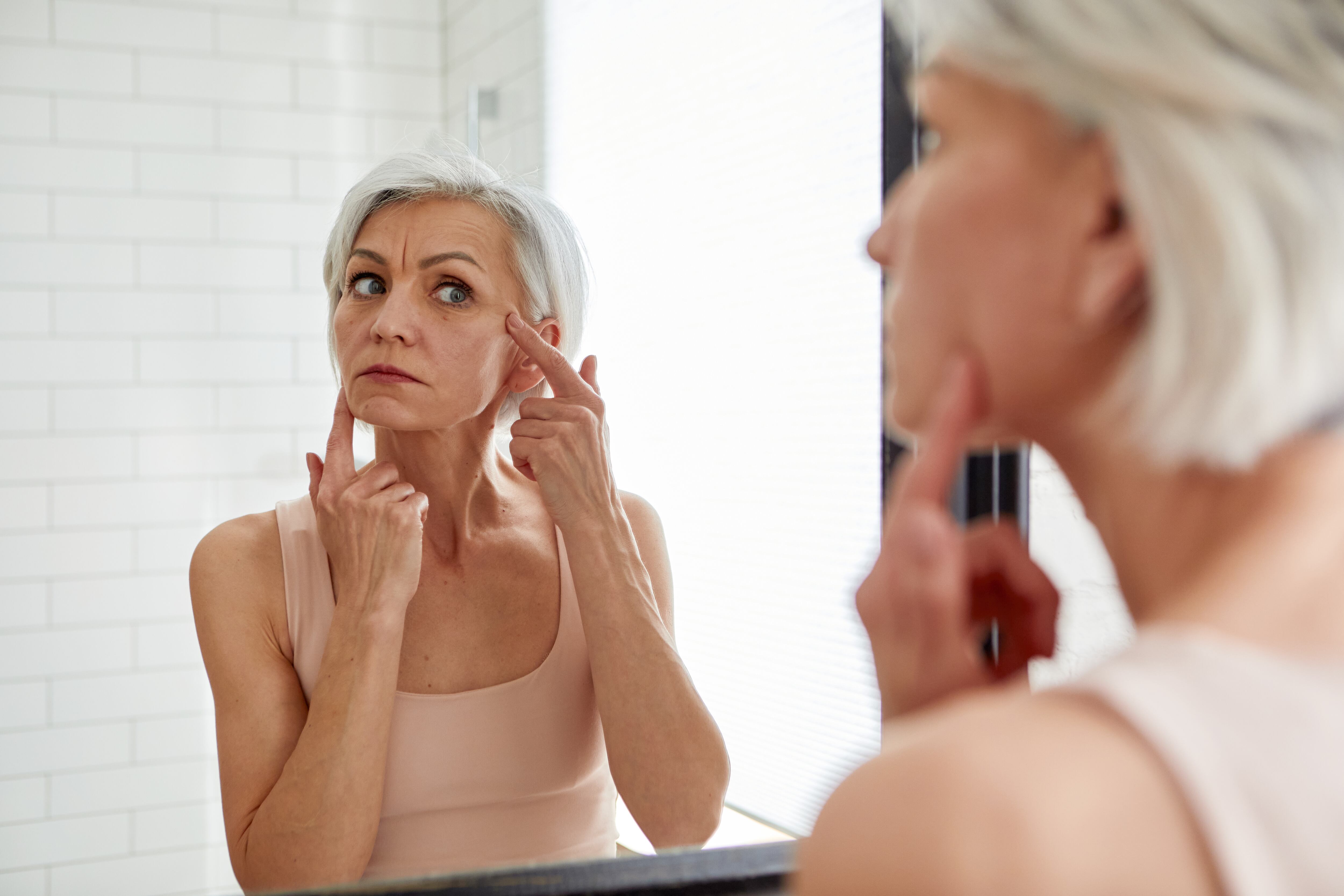 Aged gray haired female looking at herself in mirror and touching face while standing in bathroom during morning skincare routine