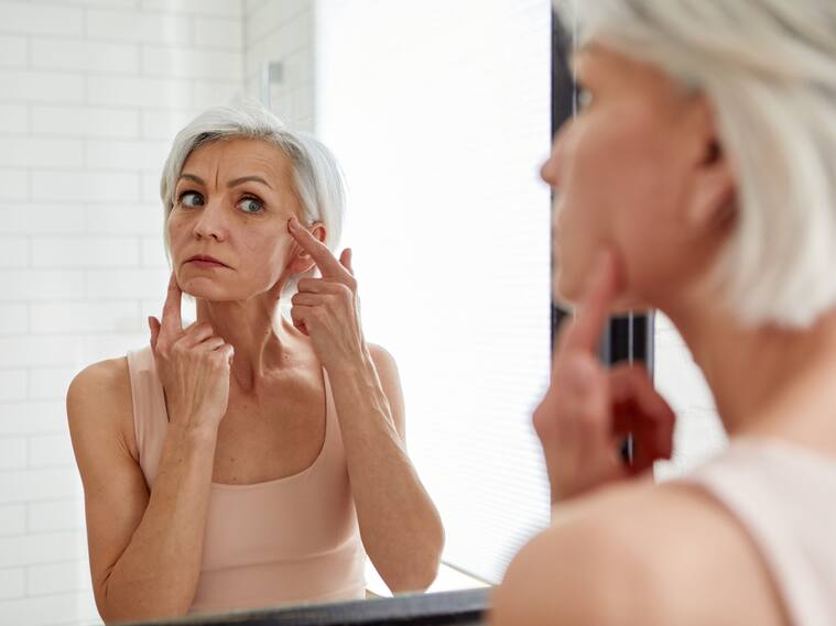 Aged gray haired female looking at herself in mirror and touching face while standing in bathroom during morning skincare routine