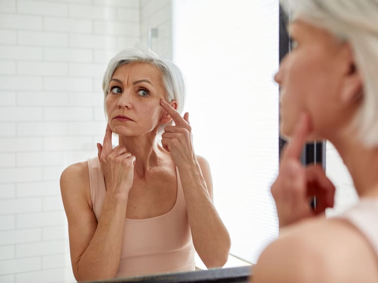 Aged gray haired female looking at herself in mirror and touching face while standing in bathroom during morning skincare routine