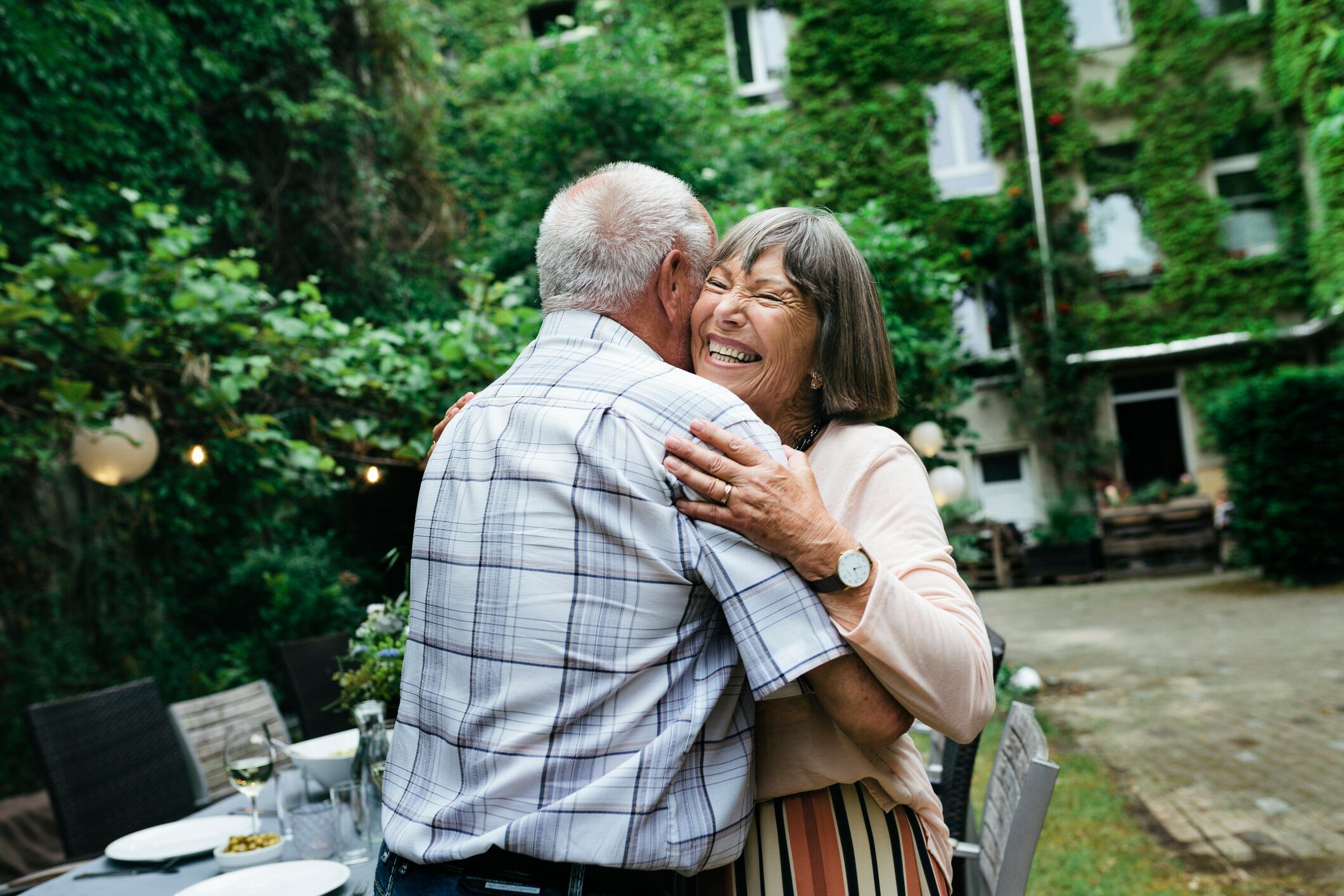 An elderly couple embracing and smiling before sitting down for a barbecue with the family in a courtyard.