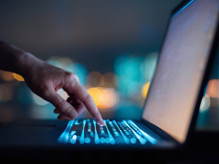 Close up of woman's hand typing on computer keyboard in the dark against colourful bokeh in background, working late on laptop at home