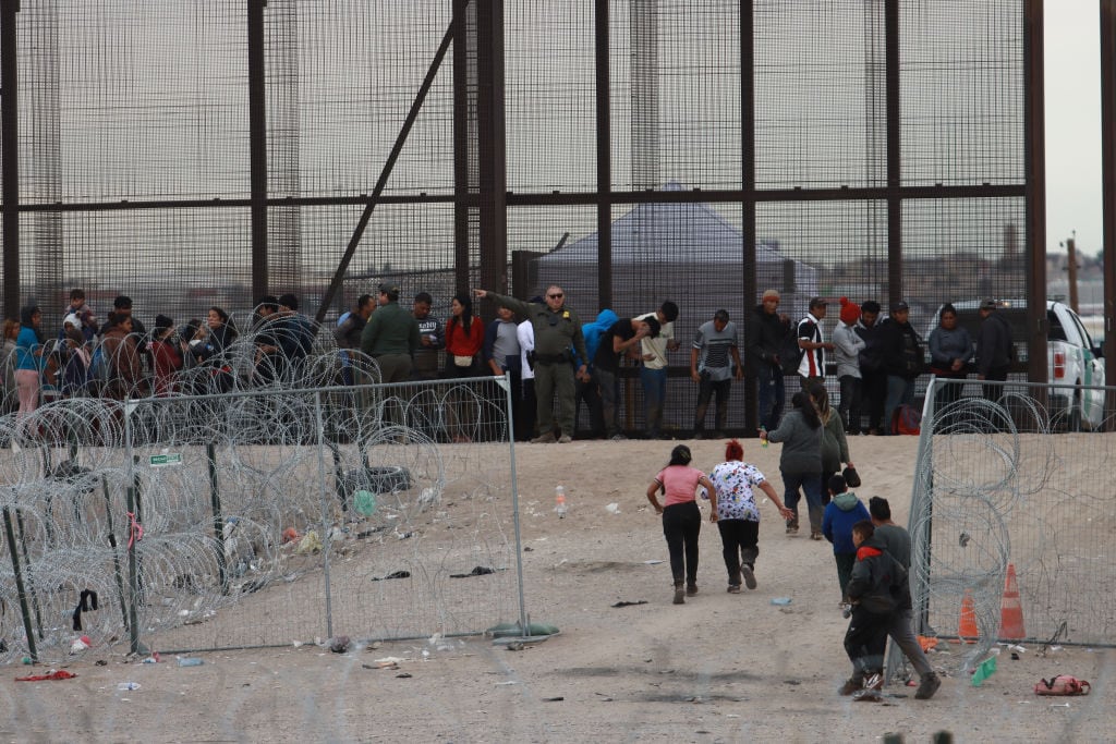 Un grupo de migrantes intentan cruzar el límite desde México hacia Estados Unidos, ante la mirada de agentes fronterizos de EEUU.