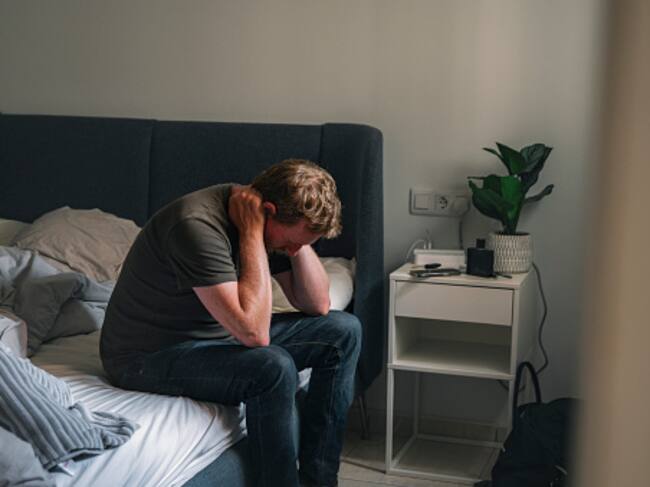 A depressed mature male sits on his bed. He clasps his hand behind his head