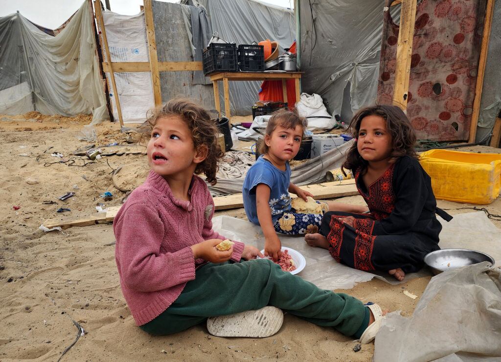 Tres niños palestinos desplazados esperan alimentos, mientras la población sufre el hambre en la Franja de Gaza en Palestina.