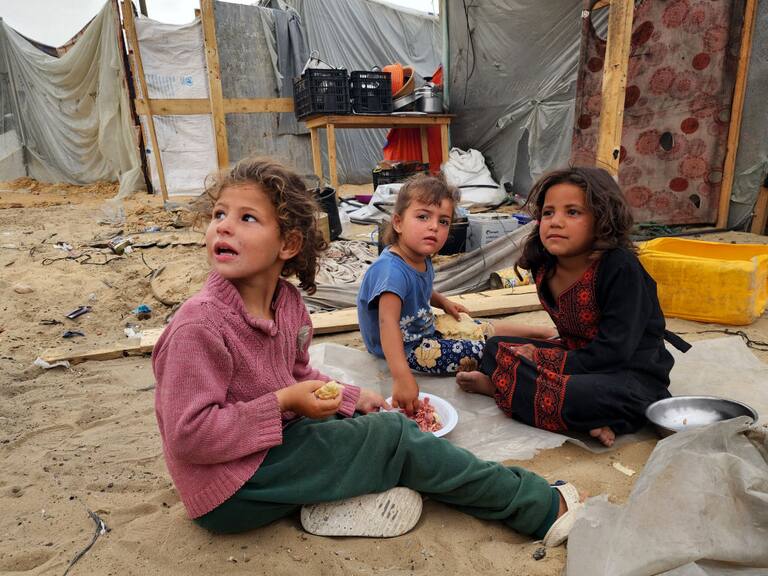 Tres niños palestinos desplazados esperan alimentos, mientras la población sufre el hambre en la Franja de Gaza en Palestina.