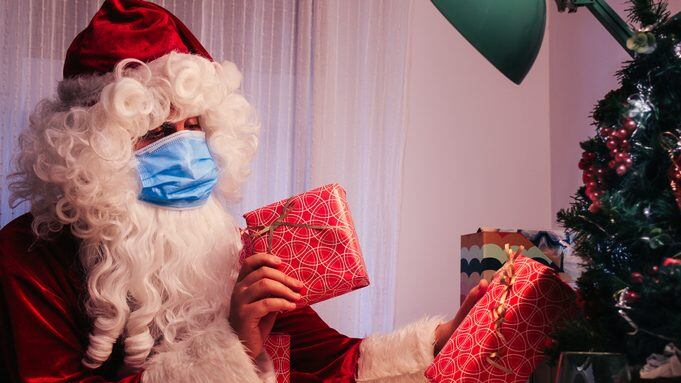 santa claus inside a house with a blue face mask, a glass of milk and cookies putting gifts on christmas tree