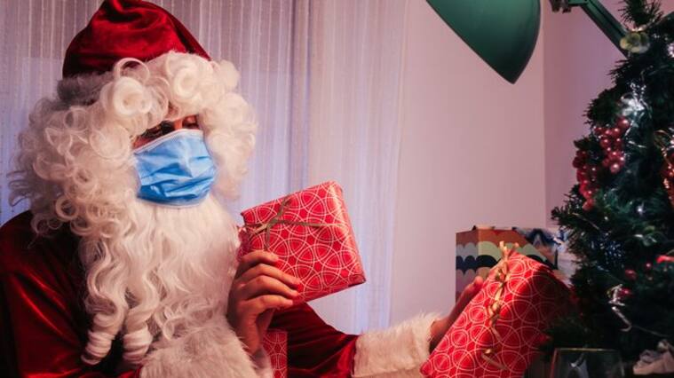 santa claus inside a house with a blue face mask, a glass of milk and cookies putting gifts on christmas tree