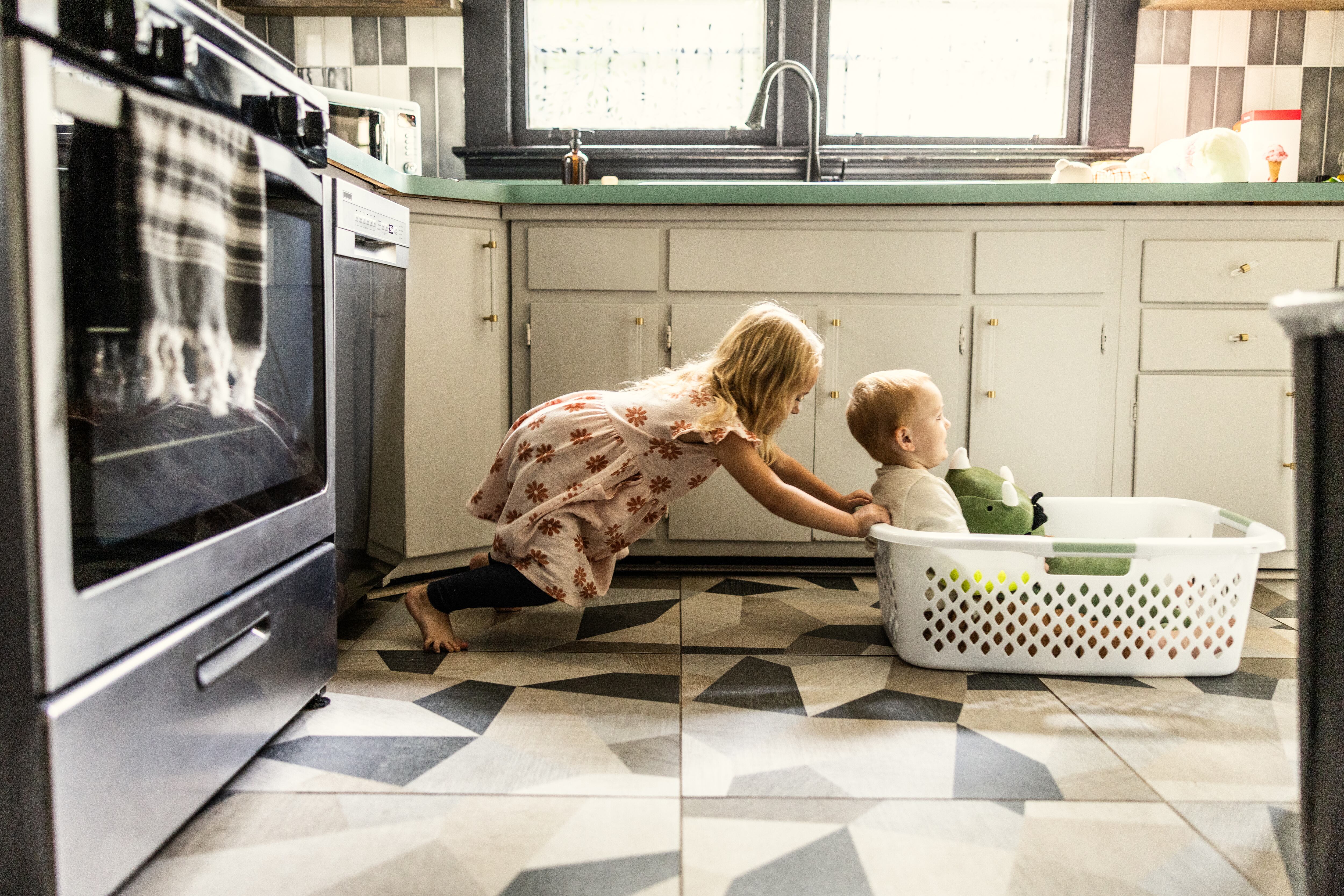 Young girl pushing toddler brother through house in laundry basket
