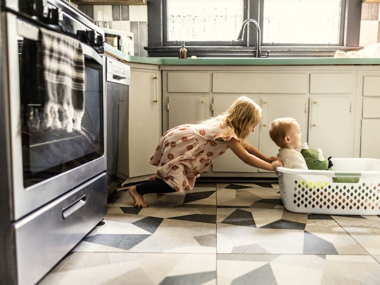 Young girl pushing toddler brother through house in laundry basket