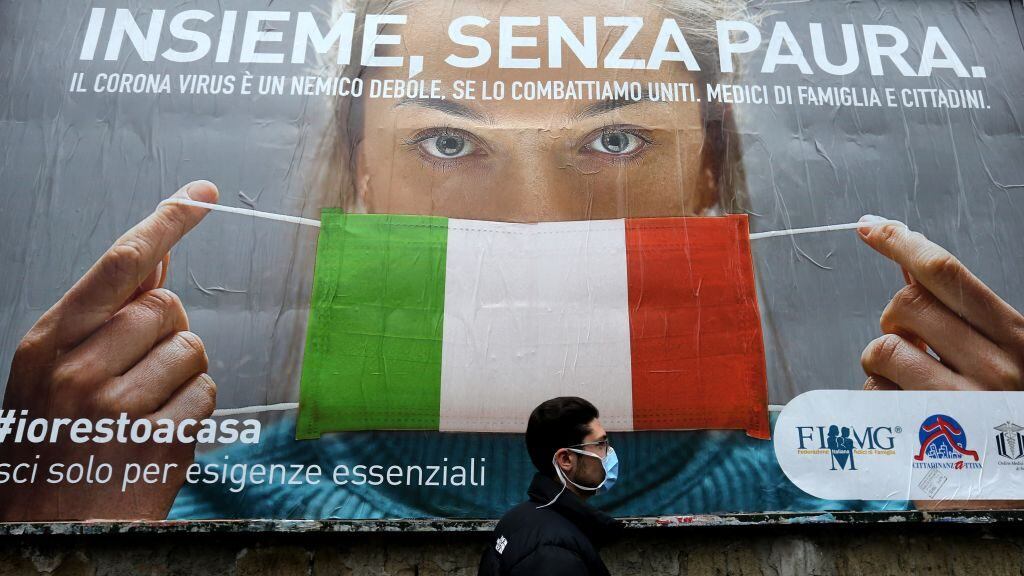 TOPSHOT - A man walks past a large billboard raising awareness to the measures   taken by the Italian government to fight against the spread of the COVID-19 (novel coronavirus) which pictures a woman wearing Italy's national flag as a facemask for protective measures and reads "All together, without fear", in the streets of Naples on March 22, 2020. - Italian Prime Minister, on March 21, ordered all non-essential companies and factories to close nationwide to stem a coronavirus pandemic that has killed 4,825 people in the country in a month. (Photo by Carlo Hermann / AFP) (Photo by CARLO HERMANN/AFP via Getty Images)