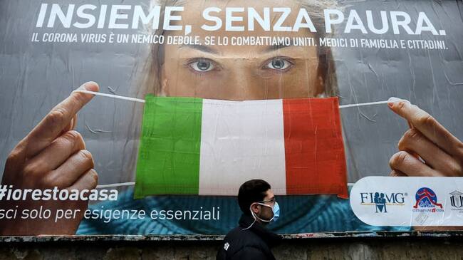 TOPSHOT - A man walks past a large billboard raising awareness to the measures taken by the Italian government to fight against the spread of the COVID-19 (novel coronavirus) which pictures a woman wearing Italy's national flag as a facemask for protective measures and reads "All together, without fear", in the streets of Naples on March 22, 2020. - Italian Prime Minister, on March 21, ordered all non-essential companies and factories to close nationwide to stem a coronavirus pandemic that has killed 4,825 people in the country in a month. (Photo by Carlo Hermann / AFP) (Photo by CARLO HERMANN/AFP via Getty Images)