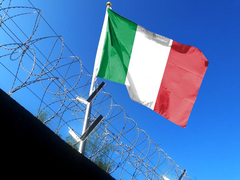 The Italian flag hangs in the cloudy sky outside the prison's barbed wire. waving in the sky