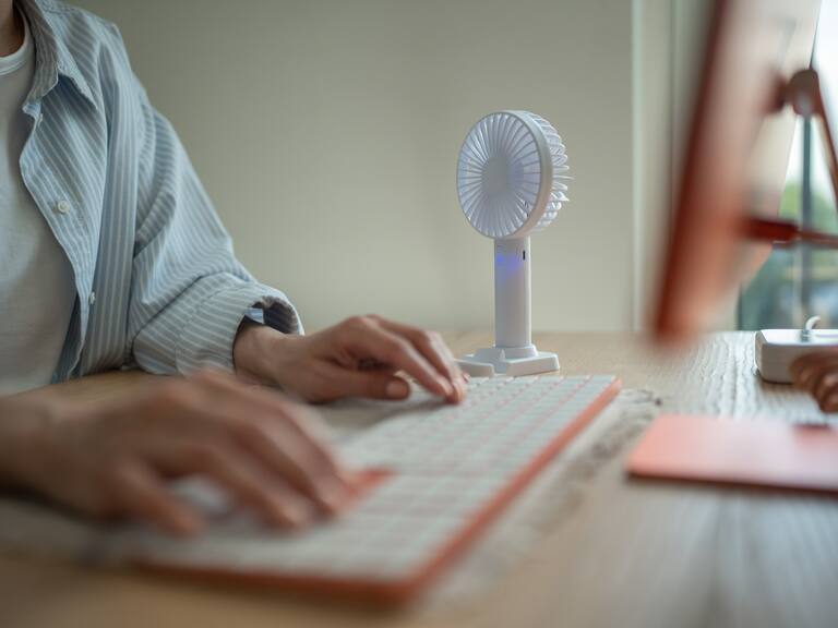 Hands of woman working typing on computer keyboard, small portable compact fan cooling air near on desk. Hot summer weather, lack of air conditioning in office, stuffiness sultriness in workplace.