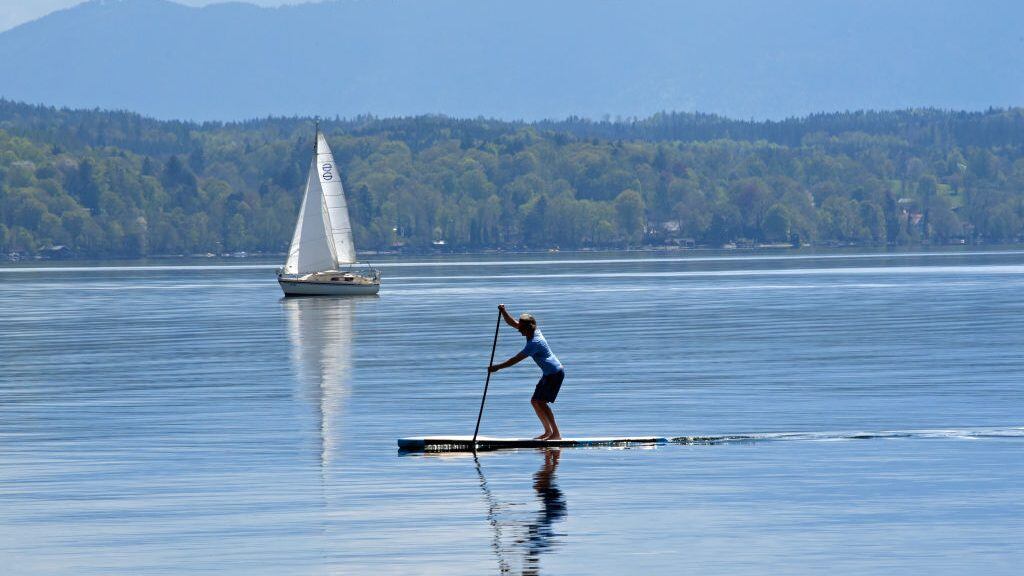 dpatop - 19 April 2020, Bavaria, Tutzing: A sailboat and a stand-up paddler drift on the unusually empty Starnberger See. Due to the exit restrictions of the Corona - Pandemic, no excursionists can reach the otherwise besieged lake despite early summer temperatures and sunshine. Photo: Ursula Düren/dpa (Photo by Ursula Düren/picture alliance via Getty Images)