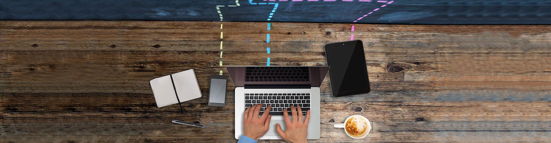 Many gadgets on wooden desk connected to the cloud. A smart phone, laptop and a tablet sharing or downloading from the cloud storage. View from above. The cloud drawn on blackboard with dashed lines going down on the desk to connect to the computers. A businessman is working on laptop