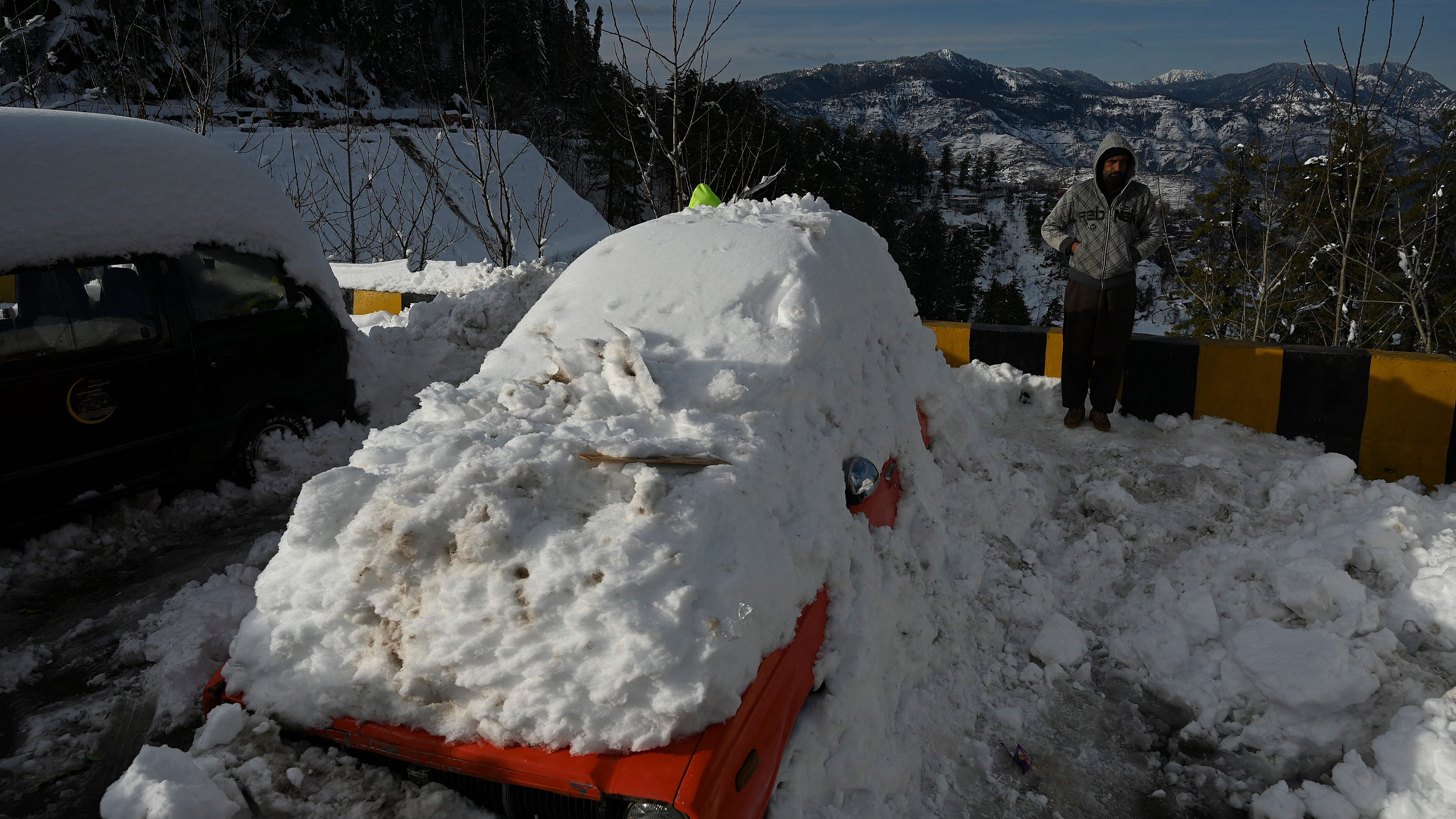 A man stands next to snow-covered card stucked on a road after a blizzard that started on January 7 and led to visitors being trapped in vehicles along the roads to the resort hill town of Murree, some 70 Kms northeast of Islamabad on January 9, 2022. - Army rescuers on January 9 cleared routes around the Pakistan hill town of Murree sheltering thousands of tourists after 22 people died in vehicles trapped by heavy snow. (Photo by Aamir QURESHI / AFP) (Photo by AAMIR QURESHI/AFP via Getty Images)
