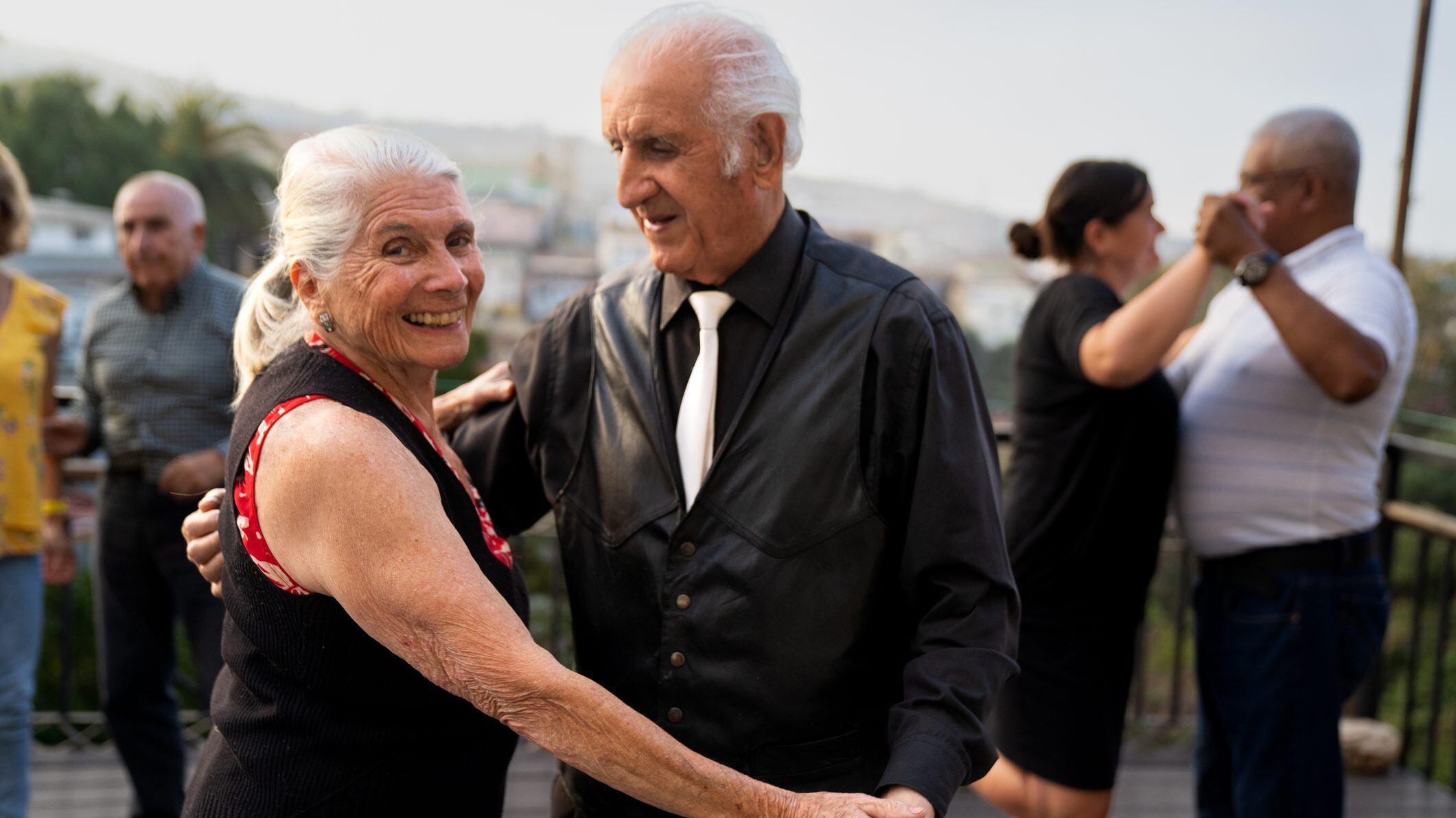 Cheerful older woman gives a big smile to the camera while in the middle of dancing with male partner in front of stunning cityscape backdrop on warm sunny day in Valparaíso, Chile