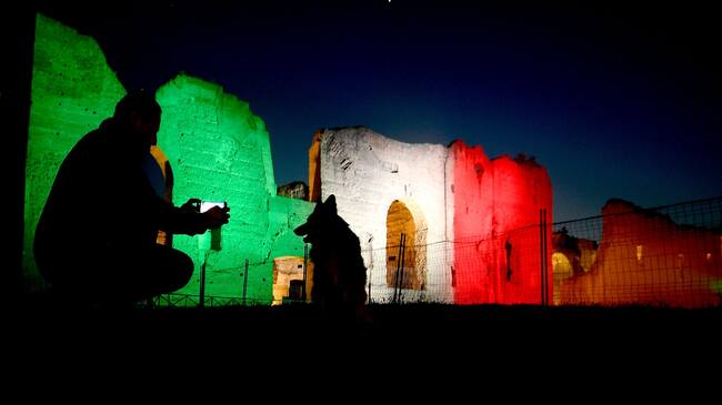 A man walks his dog by Rome's ancient Baths of Caracalla (Terme di Caracalla) lit with the colors of the Italian flag on April 10, 2020 during the country's lockdown aimed at curbing the spread of the COVID-19 infection, caused by the novel coronavirus. (Photo by Filippo MONTEFORTE / AFP) (Photo by FILIPPO MONTEFORTE/AFP via Getty Images)