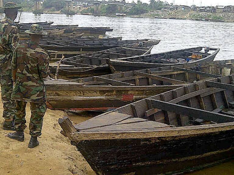 Militares de Nigeria observan las embarcaciones livianas en la orilla de un gran río.