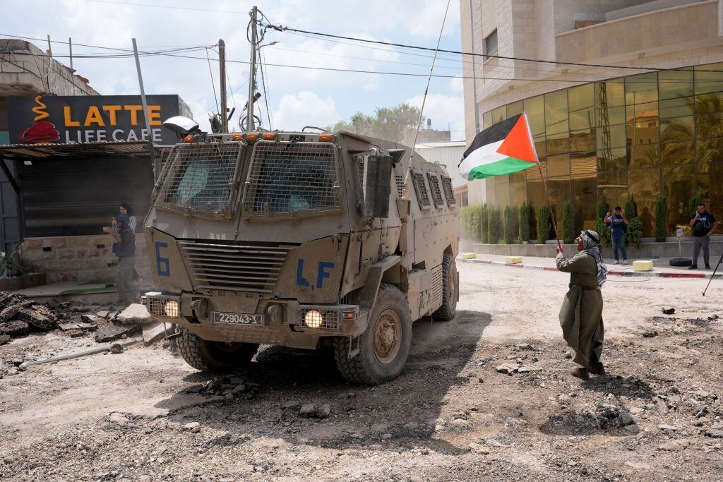 Un hombre coloca una bandera de Palestina frente a un vehículo blindado del Ejército de Israel que avanza por Cisjordania.