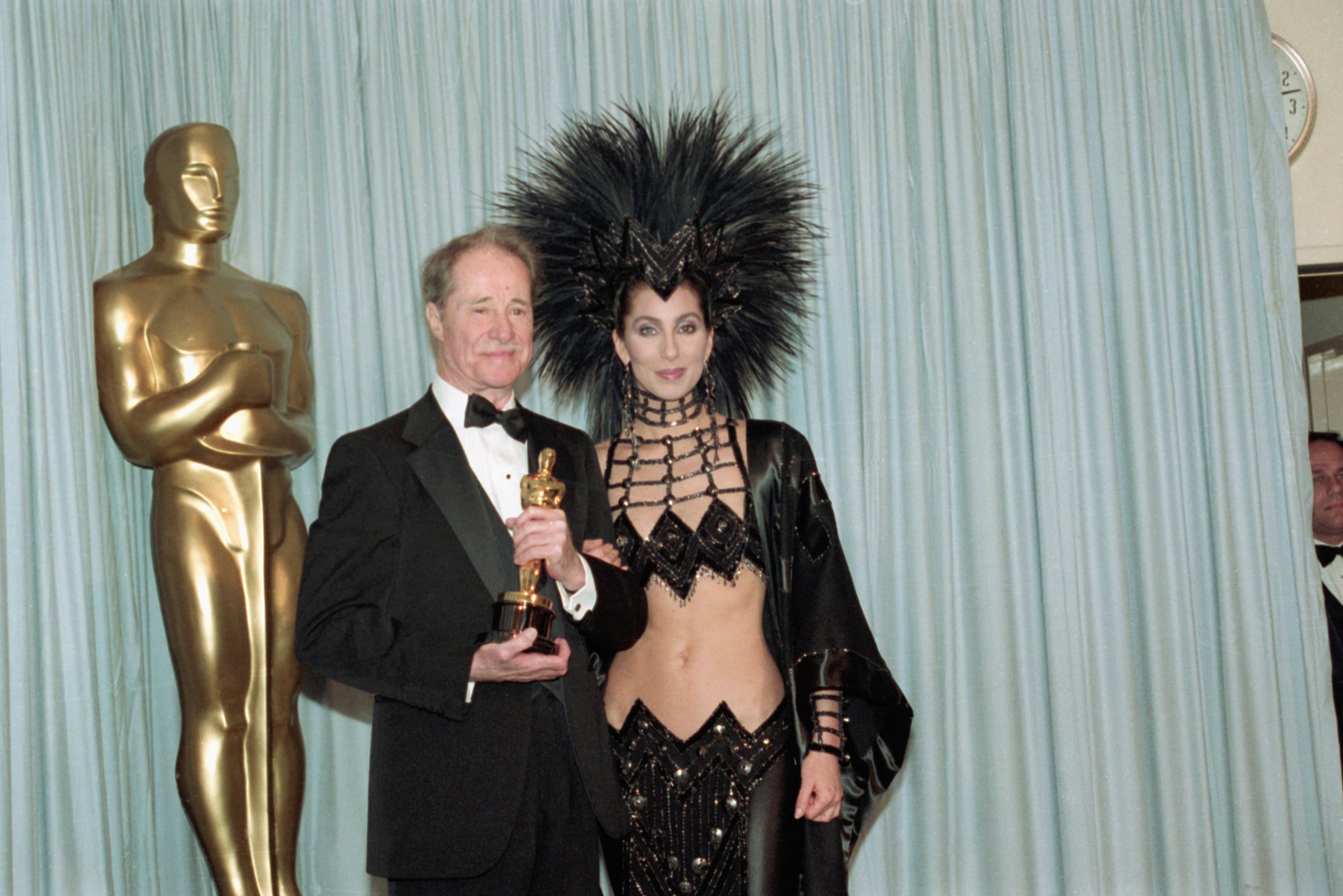 Don Ameche holding the Best Supporting Actor Oscar award for his role in Cocoon, backstage at the Academy Awards with award presenter Cher, who is wearing an unusual Bob Mackie evening gown with a feather headdress.