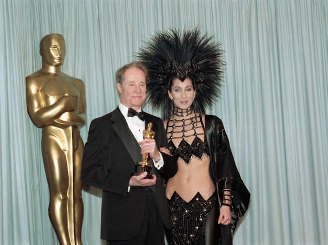 Don Ameche holding the Best Supporting Actor Oscar award for his role in Cocoon, backstage at the Academy Awards with award presenter Cher, who is wearing an unusual Bob Mackie evening gown with a feather headdress.