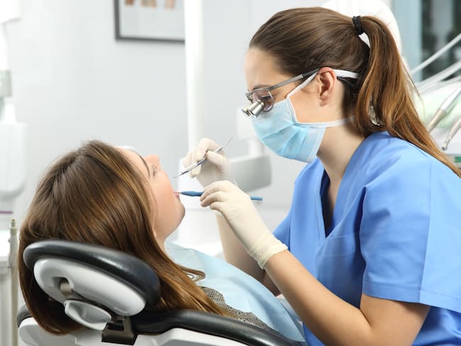 Dentist wearing eyeglasses gloves and mask examining a patient teeth with a dental probe and a mirror in a clinic box with equipment in the background
