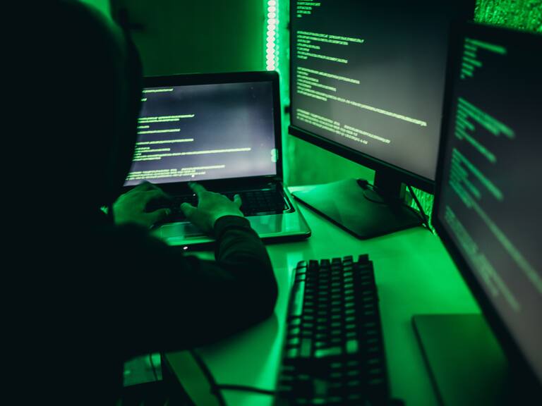 Male hacker in a black hoodie working on a computer and a laptop.