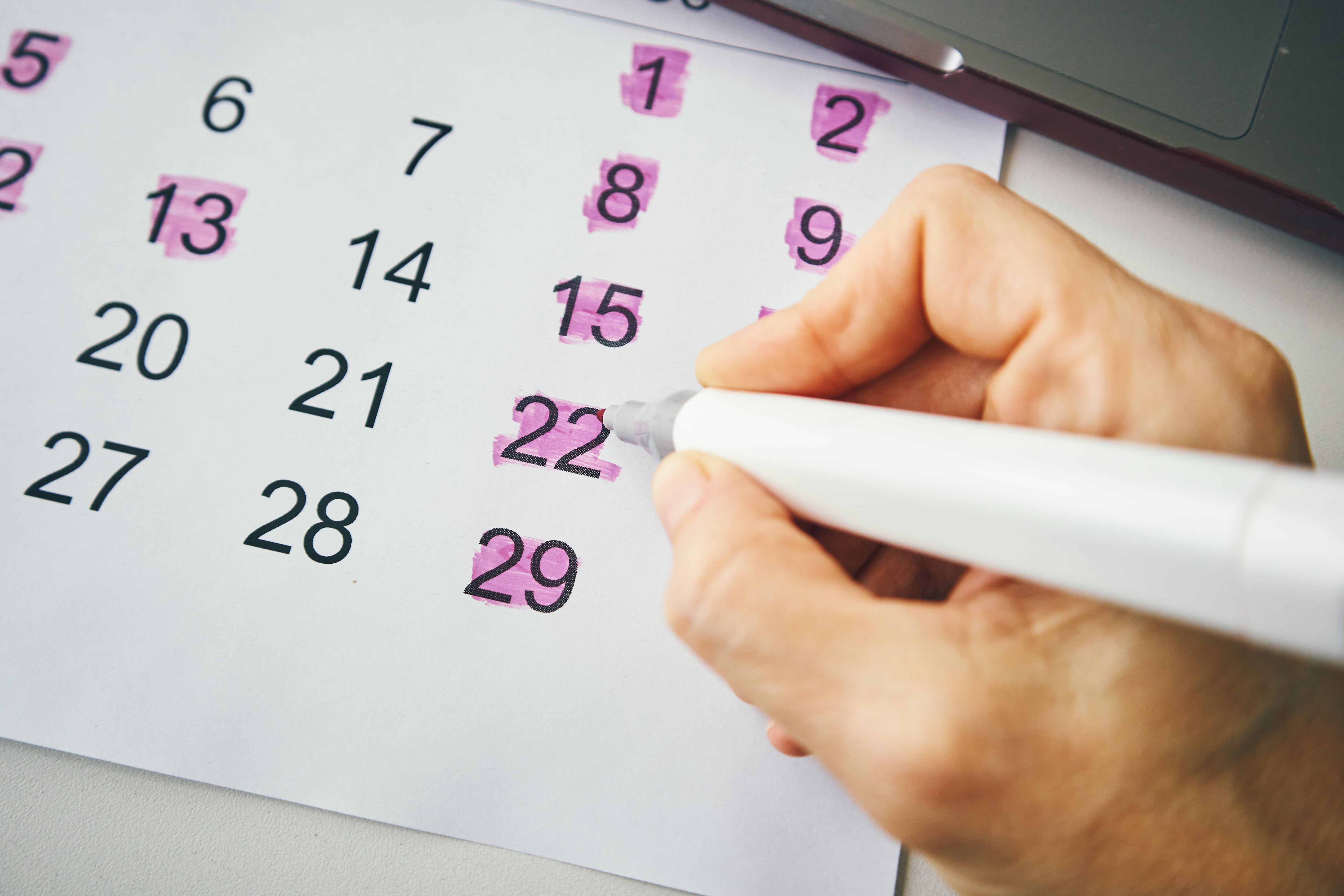 A close-up of a hand using a marker to highlight specific dates on a paper calendar.