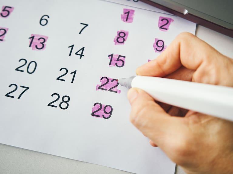 A close-up of a hand using a marker to highlight specific dates on a paper calendar.