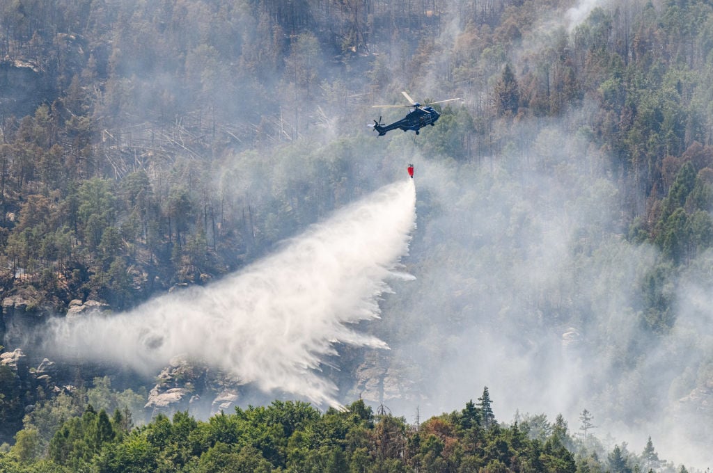 28 July 2022, Saxony, Schmilka: A helicopter of the German Federal Police flies with an external water tank to extinguish a forest fire in the Saxon Switzerland National Park. Photo: Robert Michael/dpa (Photo by Robert Michael/picture alliance via Getty Images)