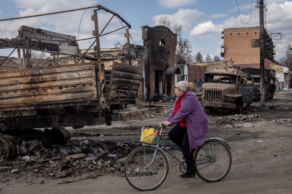 Mujer en bicicleta pasa ante vehículos destruidos por ataques en Trostyanets