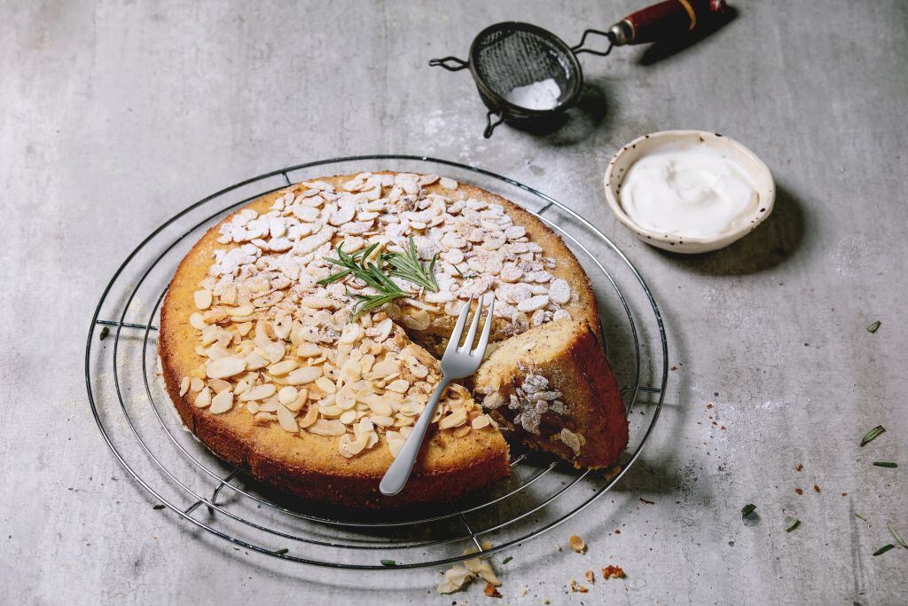 Gluten free almond rosemary homemade cake decorated by almond and sugar powder on cooling rack, serving with sour cream, fork and vintage sieve over grey texture background.. (Photo by: Natasha Breen/REDA/Universal Images Group via Getty Images)